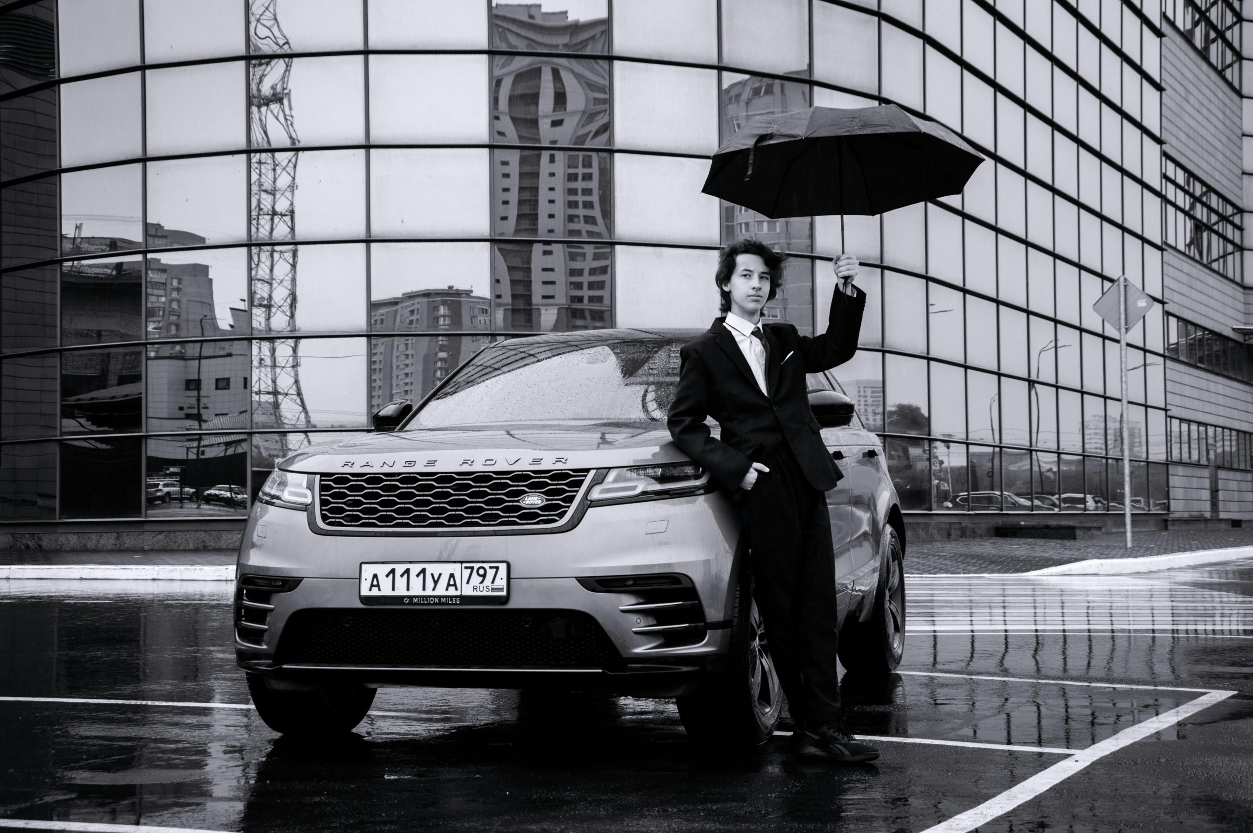 A man in a suit stands by a car with an umbrella in a rainy cityscape.